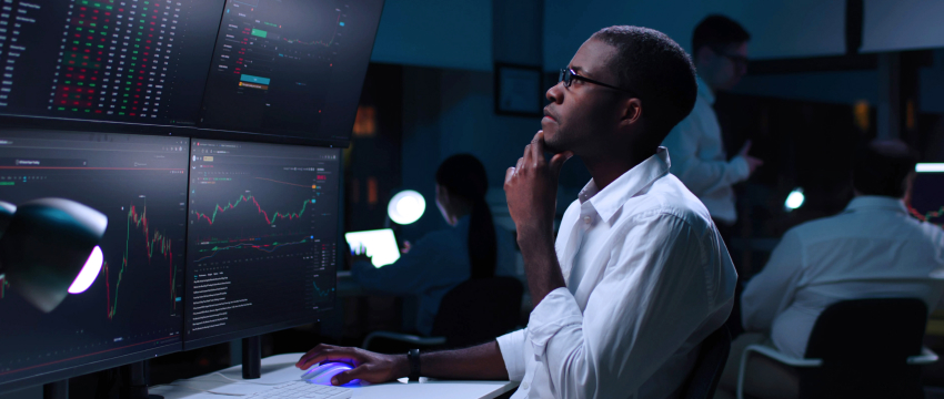 A man seated at a desk with several monitors, reviewing forex trading strategies to avoid common mistakes for success.