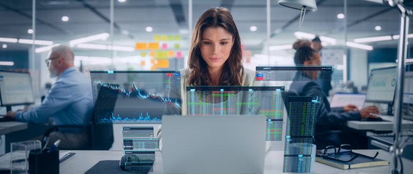 Une femme est assise à un bureau dans un bureau moderne, concentrée sur son ordinateur portable tandis que des graphiques et des courbes de trading forex sont projetés devant elle. D’autres personnes travaillent à des bureaux en arrière-plan.
