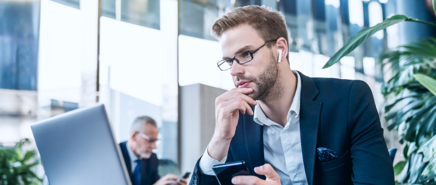 A forex trader, a man in a suit, wearing glasses and earbuds, sits thoughtfully with a phone before a laptop. Another person is blurred in the background. Office setting, focused mood trading market